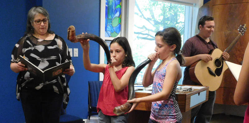 Two young girls playing horns (shofar) with Rabbi Dressler. Man with guitar in background.